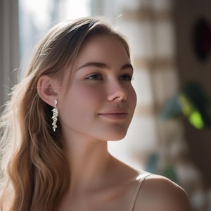 以下が含まれることがあります： 女性が揺れるイヤリングを着用しています。イヤリングはシルバー色で、葉の連なりをデザインしています。女性は長い金髪で、横を向いています。背景はぼやけており、自然光が差し込んでいます。