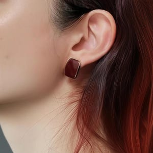 Könnte beinhalten: Nahaufnahme eines weinroten, rechteckigen Ohrrings mit goldfarbenem Rand. Der Ohrring wird am Ohr einer Person mit dunklem Haar getragen. Der Ohrring hat eine glatte, polierte Oberfläche.
