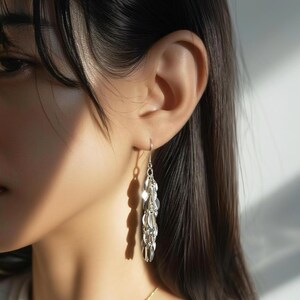 以下が含まれることがあります： フックから複数の楕円形のチャームがぶら下がっているシルバーのドロップイヤリング。イヤリングは耳に装着されており、光が光沢のある金属に反射しています。シンプルでありながらエレガントなアクセサリーです。