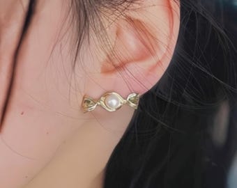 Könnte beinhalten: Goldfarbener Ohrstecker mit einer kleinen, runden Perle in der Mitte. Der Ohrring hat ein zartes, verschlungenes Design. Der Ohrring wird an einem Ohr gezeigt, wobei der Fokus auf dem Schmuck liegt.