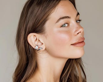 Puede incluir: Un pendiente de oreja de zorro plateado con un acento de cristal brillante. El pendiente se envuelve alrededor de la oreja, con un diseño de cabeza y cola de zorro. La joya se lleva en la oreja de una persona con cabello castaño largo.