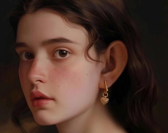 Puede incluir: Retrato de una persona con cabello castaño y ojos marrones. La persona lleva un pendiente dorado con un colgante en forma de corazón. La piel tiene una tez clara con un ligero rubor en las mejillas y los labios. El fondo es oscuro.