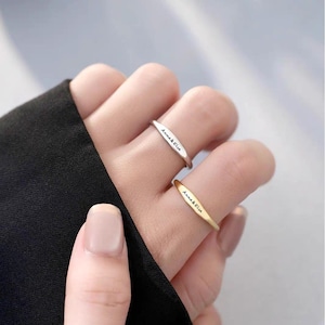 Può includere: Due anelli sottili in oro e argento con l'iscrizione "Anna & Ella" su di essi. Gli anelli sono indossati al dito di una persona.
