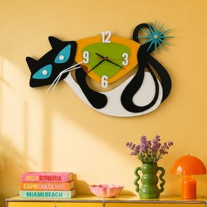 May include: A wall clock shaped like a stylized black and white cat with blue eyes and a green triangular face featuring white numbers 3, 6, 9, and 12. The clock has black hands and a blue starburst detail on the cat's tail, mounted on a yellow wall.