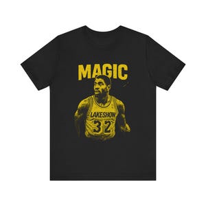 May include: Black t-shirt featuring a yellow graphic of a basketball player with the word "MAGIC" above his head. The player is wearing a yellow jersey with the word "LAKESHOW" and the number "32".