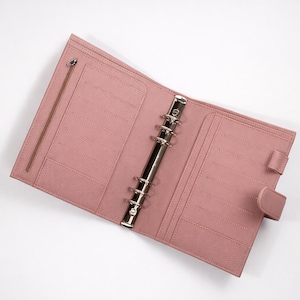 May include: Open, pink leather planner with a silver binder mechanism. The planner has multiple card slots, a zipped pocket, and a snap closure. The interior is a matching pink colour.