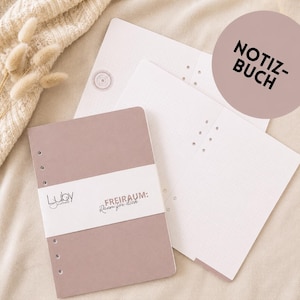 Può includere: Un set di quaderni con copertina malva e pagine bianche. La copertina presenta il testo "Luby Journals" e "FREIRAUM: Raum für dich". Le pagine del quaderno hanno un motivo a pois. È visibile anche un adesivo rotondo con la scritta "NOTIZ-BUCH".