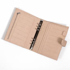 May include: Open, light tan leather planner with a silver binder mechanism. The planner has multiple card slots, a zipped pocket, and a snap closure. The interior is a matching light tan colour.