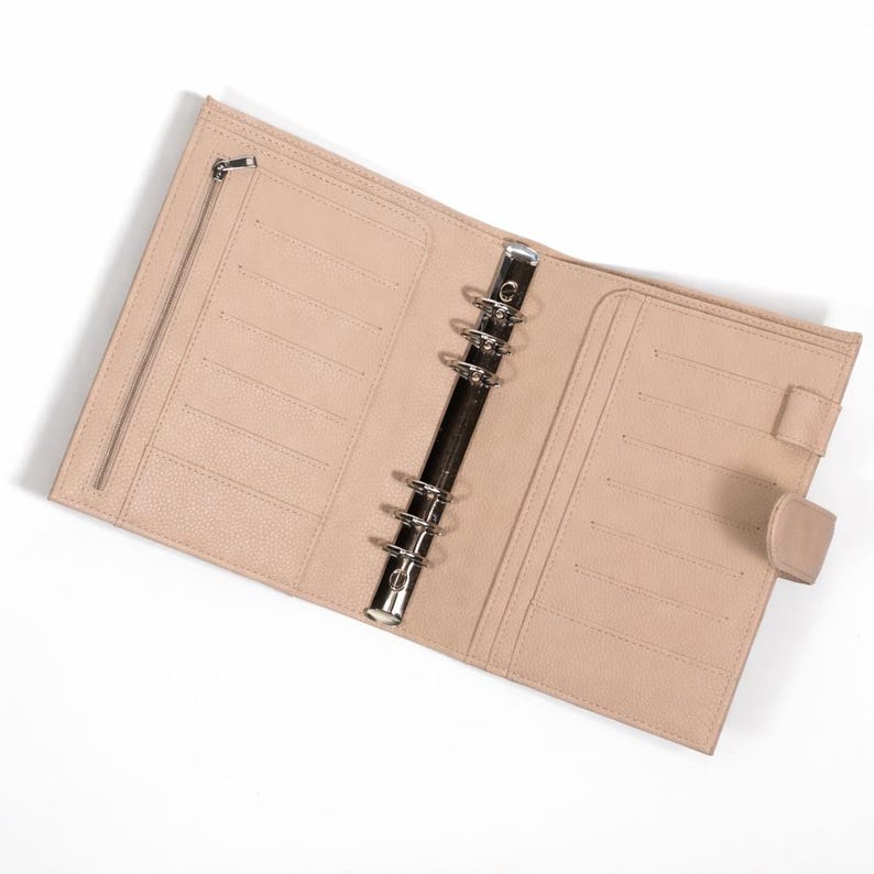 May include: Open, light tan leather planner with a silver binder mechanism. The planner has multiple card slots, a zipped pocket, and a snap closure. The interior is a matching light tan colour.