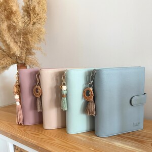 May include: Four pastel-coloured leather journals with decorative keychains featuring tassels and wooden beads. The journals are arranged on a wooden shelf, with a dried plant in the background. The journals are in shades of pink, peach, mint, and blue.