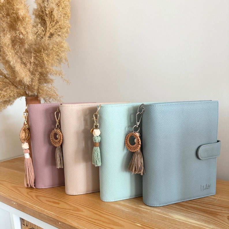 May include: Four pastel-coloured leather journals with decorative keychains featuring tassels and wooden beads. The journals are arranged on a wooden shelf, with a dried plant in the background. The journals are in shades of pink, peach, mint, and blue.