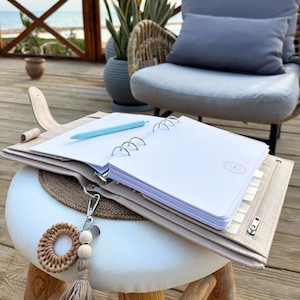 Peut inclure: Un agenda en cuir beige avec un carnet ouvert, un stylo bleu clair et un porte-clés décoratif. L'agenda contient du papier à points et une fermeture éclair. Le porte-clés a des perles en bois et un pompon.