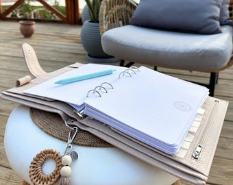 Könnte beinhalten: Ein beigefarbener Lederplaner mit einem offenen Notizbuch mit Punktraster und einem hellblauen Stift. Ein dekorativer Schlüsselanhänger mit Holzperlen und Quaste hängt am Planer. Der Planer liegt auf einem kleinen weißen Hocker auf einer Holzterrasse.
