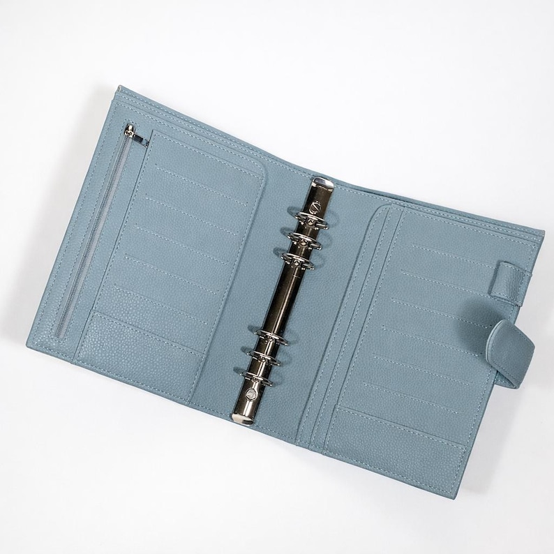 May include: Open, pale blue leather binder with a silver metal ring mechanism. The binder features multiple card slots, a zipped pocket, and a snap closure. The binder is displayed open against a white backdrop.