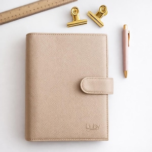 May include: A beige leather planner with a tab closure, a pink pen, two gold binder clips, and a wooden ruler are arranged on a white surface. The planner has the brand name "Luby" embossed on the lower right corner.