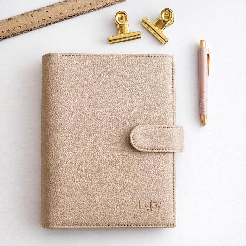 May include: A beige leather planner with a tab closure, a pink pen, two gold binder clips, and a wooden ruler are arranged on a white surface. The planner has the brand name "Luby" embossed on the lower right corner.