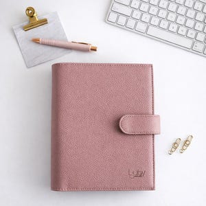 May include: A pink planner with a textured finish and a clasp. A gold pen, paper clip, and clipboard sit on a white surface. A white keyboard is in the background. The planner has the text "Luby Journals".