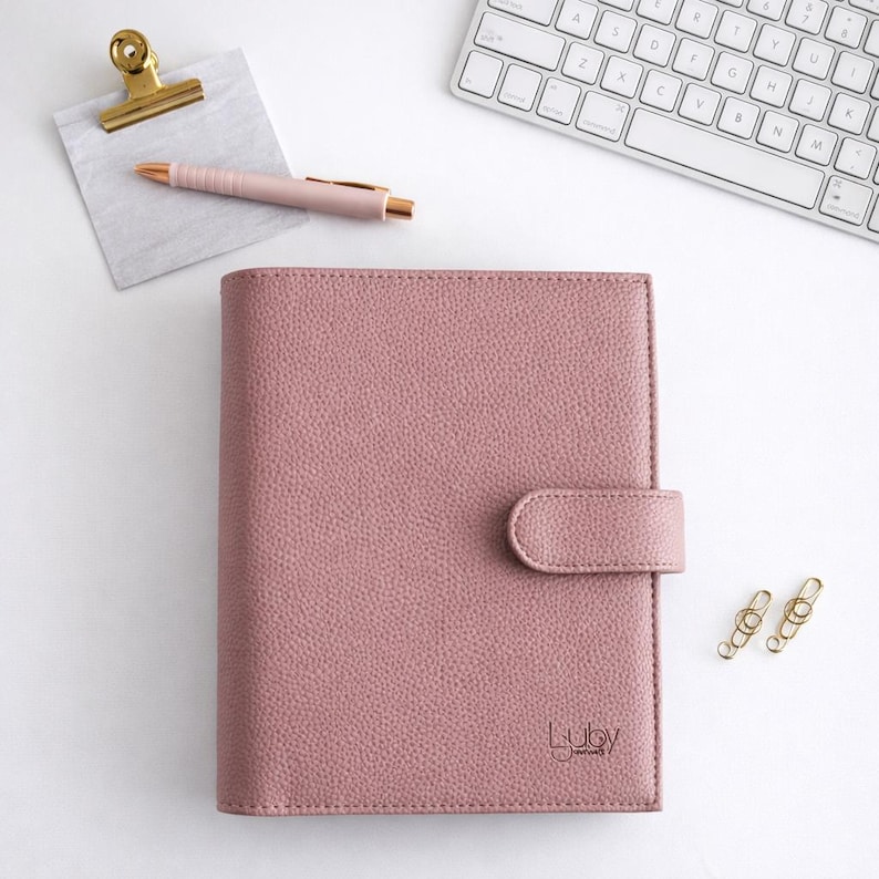 May include: A pink planner with a textured finish and a clasp. A gold pen, paper clip, and clipboard sit on a white surface. A white keyboard is in the background. The planner has the text "Luby Journals".