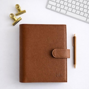 May include: A brown leather journal with a matching pen and gold-coloured binder clips. The journal has a textured surface and a clasp closure. A white keyboard is in the background. The journal has the word "Luby" printed on the bottom right.