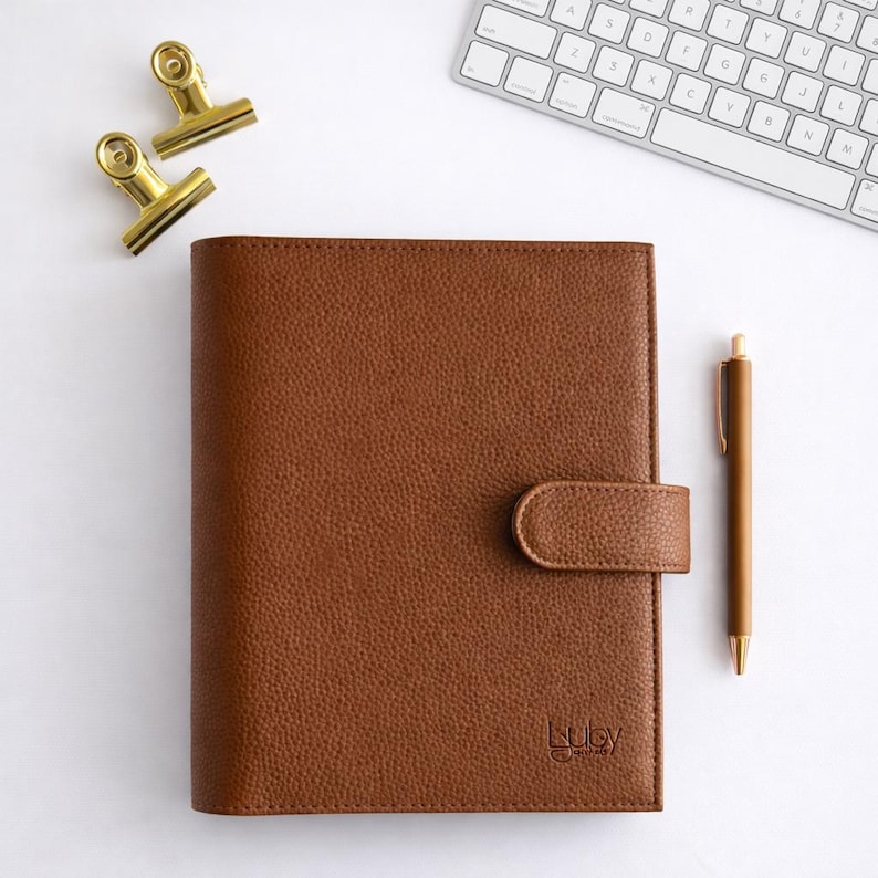 May include: A brown leather journal with a matching pen and gold-coloured binder clips. The journal has a textured surface and a clasp closure. A white keyboard is in the background. The journal has the word "Luby" printed on the bottom right.