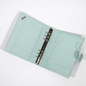 May include: Open, pale blue leather-effect binder with a silver metal ring mechanism. The binder features multiple card slots, a zipped pocket, and a tab closure. The interior is a solid, pale blue colour.