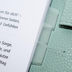 Może przedstawiać: Zbliżenie na planer z przezroczystymi plastikowymi przegrodami. Planer ma jasną, miętową skórzaną okładkę. Na białych stronach widoczny jest tekst w języku niemieckim, w tym słowa "um für dich" i "Meerer Seiten".