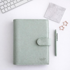May include: A pale green journal with a matching pen and star-shaped paper clips. The journal has a textured surface, a clasp closure, and the words "Luly Journal" printed on the bottom right corner.