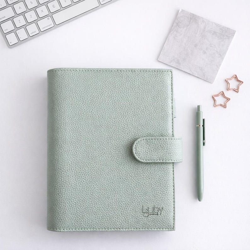 May include: A pale green journal with a matching pen and star-shaped paper clips. The journal has a textured surface, a clasp closure, and the words "Luly Journal" printed on the bottom right corner.