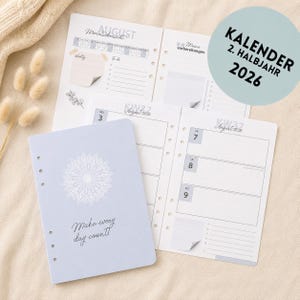 Può includere: Un'agenda azzurra con un motivo floreale bianco e la scritta "Make every day count!" è aperta per visualizzare le pagine del calendario settimanale e mensile. Le pagine del calendario sono datate agosto 2026. Un adesivo circolare recita "KALENDER 2. HALBJAHR 2026".