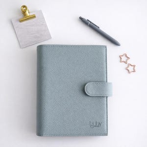 May include: A pale blue leather journal with a clasp, a grey pen, a gold paper clip, and two star-shaped paper clips. The journal has the text "luby journals" printed on the bottom right corner.