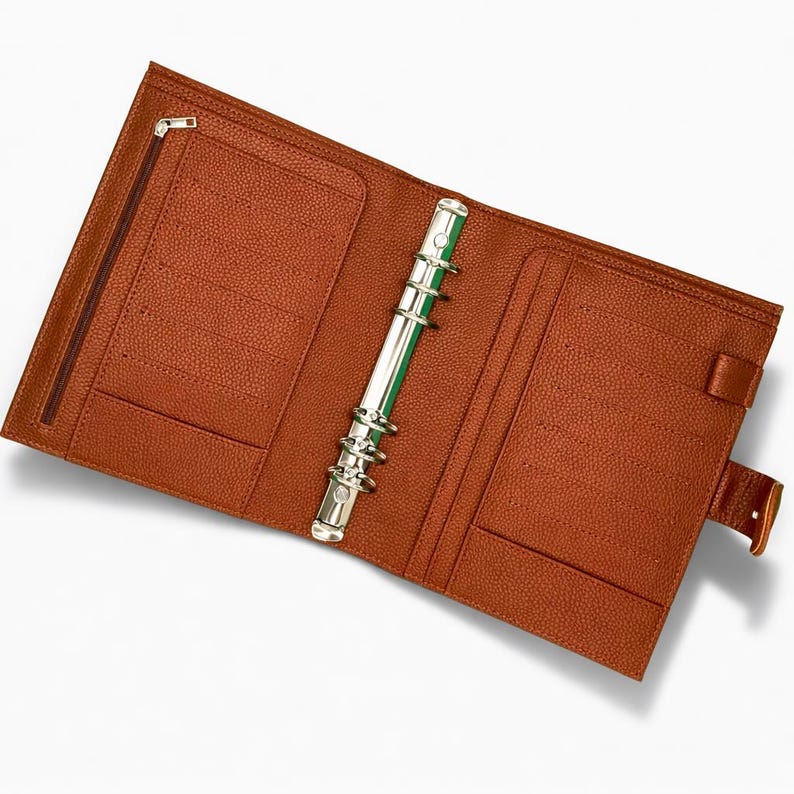 May include: Open tan leather binder with a silver metal ring mechanism. The binder has multiple pockets, a zipped compartment, and a snap closure. The interior has a textured surface.