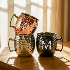 Puede incluir: Tres tazas Moscow Mule personalizadas apiladas sobre una superficie de madera. La taza superior es de cobre con un asa verde y la letra "P". La taza del medio es plateada con la letra "R". La taza inferior es negra con la letra "M".
