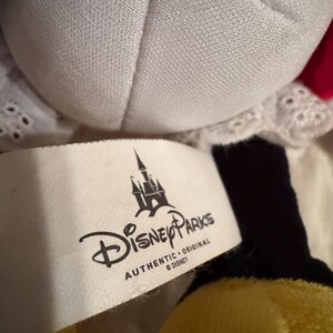 May include: A close-up of a Disney Parks plush toy. The toy has a white collar with lace trim, a red bow, and black and yellow accents. A white tag displays the Disney Parks logo and the words "AUTHENTIC ORIGINAL DISNEY".