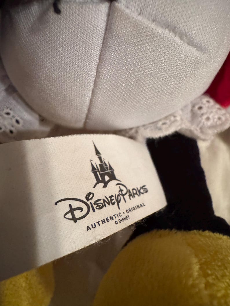 May include: A close-up of a Disney Parks plush toy. The toy has a white collar with lace trim, a red bow, and black and yellow accents. A white tag displays the Disney Parks logo and the words "AUTHENTIC ORIGINAL DISNEY".