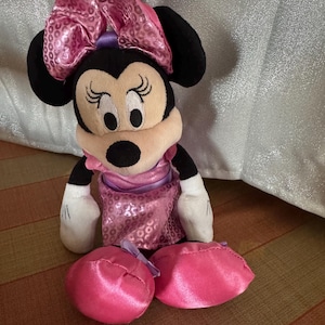 May include: A Minnie Mouse plush toy with a large pink sequined bow, black ears, and a pink dress. The toy has white gloves and two pink satin shoes. The plush is sitting on a striped surface.