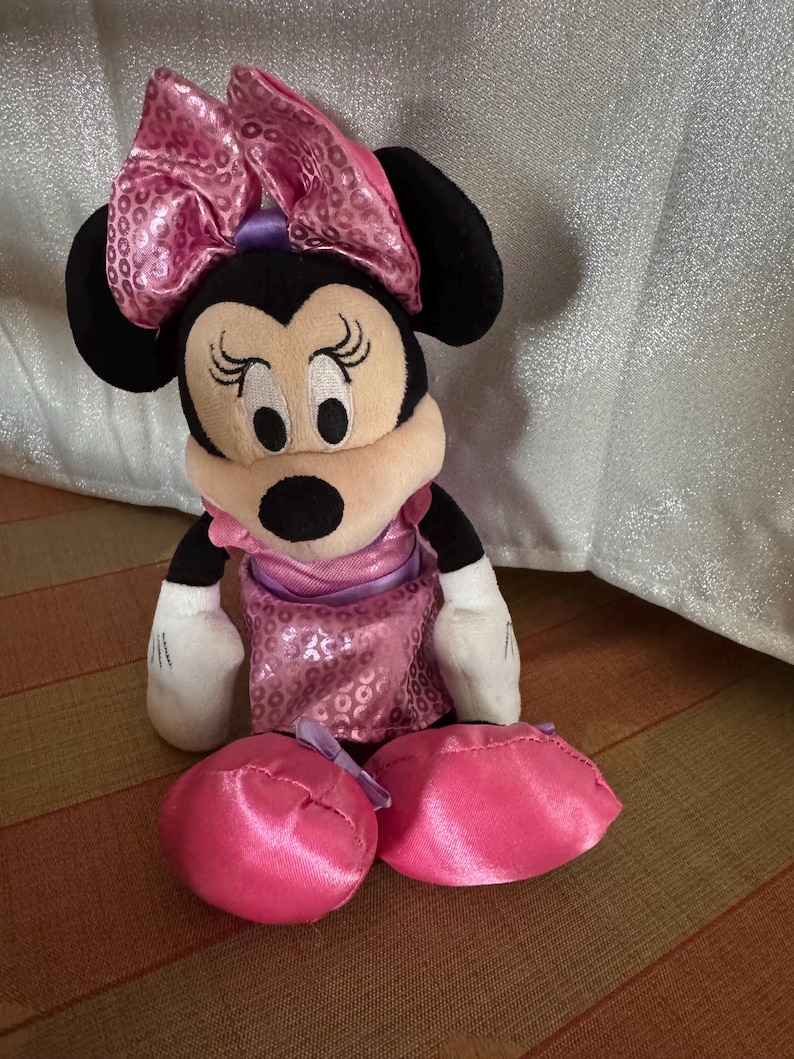 May include: A Minnie Mouse plush toy with a large pink sequined bow, black ears, and a pink dress. The toy has white gloves and two pink satin shoes. The plush is sitting on a striped surface.