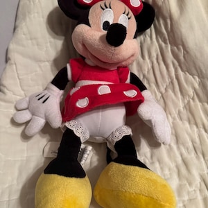 May include: A plush Minnie Mouse doll, featuring a red dress with white polka dots, a matching bow, and yellow shoes. The doll has black ears, arms, and legs, and white gloves. The doll is a classic Disney character.