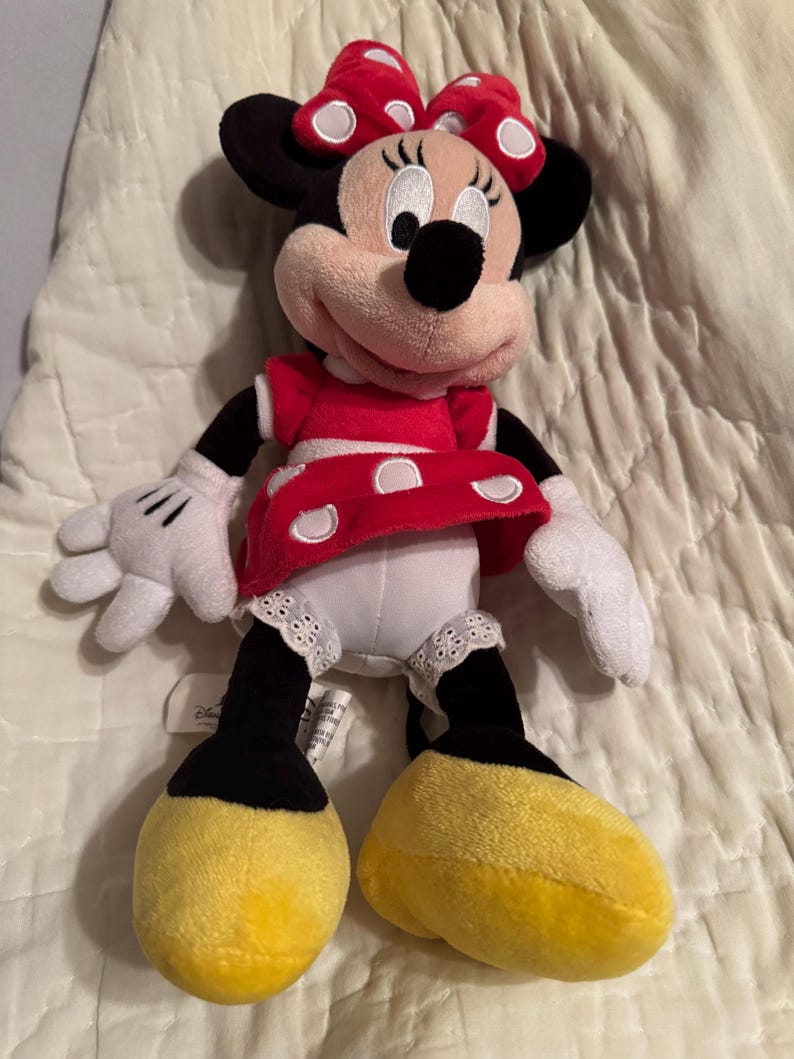 May include: A plush Minnie Mouse doll, featuring a red dress with white polka dots, a matching bow, and yellow shoes. The doll has black ears, arms, and legs, and white gloves. The doll is a classic Disney character.