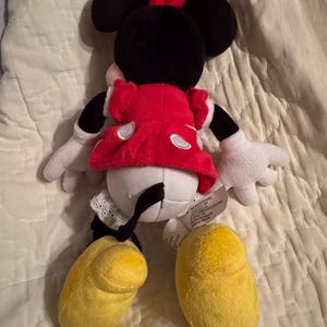 May include: A plush Minnie Mouse doll, viewed from the back, with a red dress, white gloves, and yellow shoes. The doll has black ears and a red bow. The doll is on a white surface.