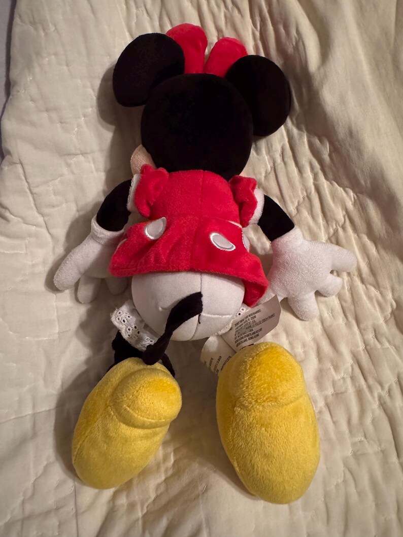May include: A plush Minnie Mouse doll, viewed from the back, with a red dress, white gloves, and yellow shoes. The doll has black ears and a red bow. The doll is on a white surface.
