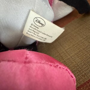 May include: Close-up of a Disney plush toy with a pink dress and black ears. A white tag with the Disney logo and text is visible. The tag includes the manufacturer's name and address.