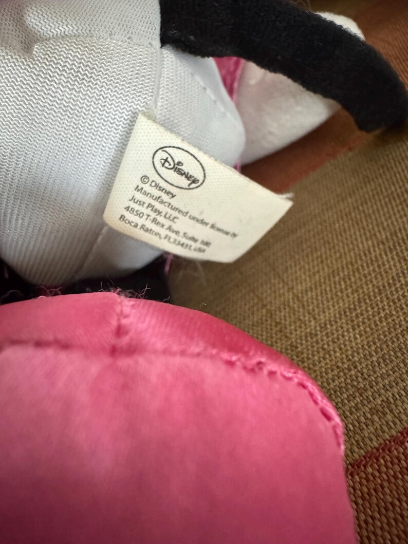 May include: Close-up of a Disney plush toy with a pink dress and black ears. A white tag with the Disney logo and text is visible. The tag includes the manufacturer's name and address.