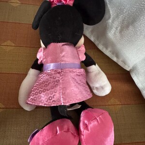 May include: A Minnie Mouse soft toy, seen from behind, has a black head with large ears and a pink bow. She is dressed in a pink dress with a purple belt and pink shoes. The doll has white gloves and is made of soft fabric.