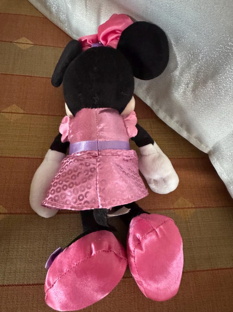 May include: A Minnie Mouse soft toy, seen from behind, has a black head with large ears and a pink bow. She is dressed in a pink dress with a purple belt and pink shoes. The doll has white gloves and is made of soft fabric.
