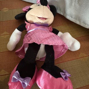 May include: A Minnie Mouse soft toy. The toy features a beige face, black ears, and a pink dress with a matching bow. She wears pink satin shoes with purple bows. The toy is on a patterned surface.