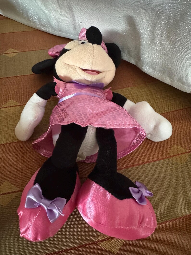 May include: A Minnie Mouse soft toy. The toy features a beige face, black ears, and a pink dress with a matching bow. She wears pink satin shoes with purple bows. The toy is on a patterned surface.