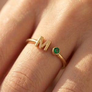 May include: A gold open-style ring featuring a capital letter M on one end and a small, round, bezel-set green gemstone on the other end. The band is thin and polished, creating a simple, continuous loop design that rests on a finger.