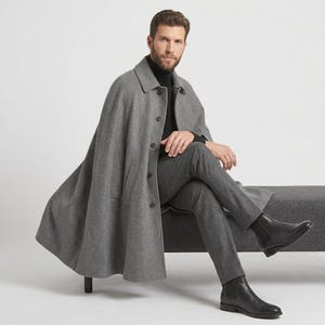 May include: A grey wool-blend cape coat featuring a classic collar, a front button placket with five visible buttons, and side slits for arm movement. The garment is worn over a black turtleneck sweater, dark grey trousers, and black leather boots.