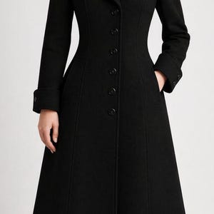 May include: A long, black, tailored coat featuring a structured collar, long sleeves with cuffed wrists, and a single-breasted front closure with six visible black buttons. The coat has a fitted waistline and a flared, floor-length skirt design.
