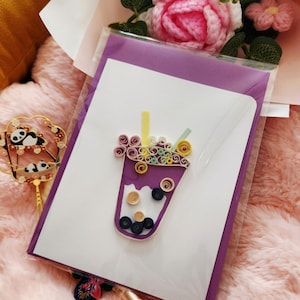 May include: A handmade greeting card featuring a purple bubble tea design made with paper quilling. The card is inside a clear plastic sleeve, next to a bouquet of pink and green flowers and a decorative panda fan.
